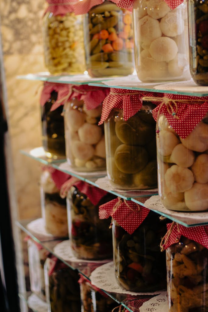 Homemade pickled vegetables in jars with red cloth caps on rustic shelves.